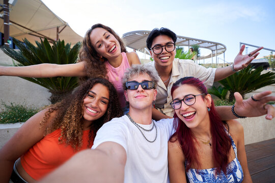 Five cheerful diverse friends taking a selfie photo while making funny faces, gathering together outdoors. Multiracial group of young people sharing memories on social media.