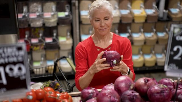 A senior woman selects fresh vegetables and fruits with a smile while shopping at a grocery store. She enjoys her time choosing food.