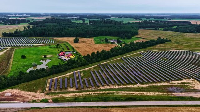 Aerial view of solar farm panels arranged in rows across a rural landscape with a small pond and a house near Tume, Tukums Municipality, Latvia.