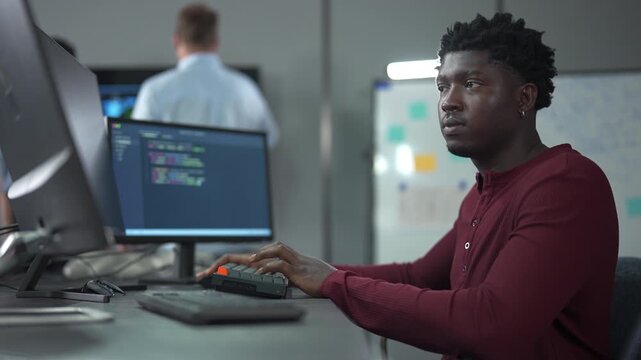 Young African male software engineer writing program code on multiple desktop screens while his colleagues working in the background in a modern office.