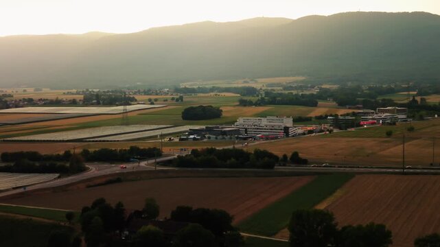 Aerial view of Rte Blanche 1 industrial complex surrounded by agricultural fields and meadows at sunset in Signy-Avenex, Vaud, Switzerland.