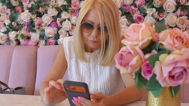 Mature blonde woman using smartphone in a pink floral cafe