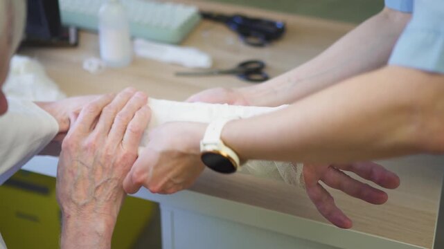 Medical staff carefully wraps a bandage around the injured hand of a senior patient in a clinic. The professional applies a cast to secure the fracture with gentle, focused care.