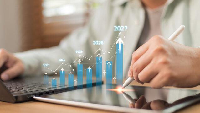 Businessman using stylus on tablet analyzing financial growth chart, pointing upward arrow towards 2027. Concept of business development, market forecast, investment strategy and future trend