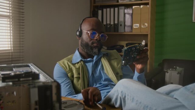 African American man wearing headphones inspecting computer graphics card at desk. Video suits IT support services, hardware repair, and technology education commercials