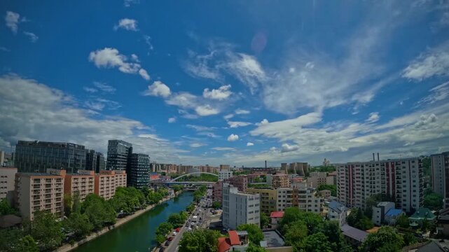Elevated wide-angle time-lapse showing fluffy white clouds moving across a vibrant blue summer sky over Bucharest, with traffic flowing like ants along the D&acirc;mbovița River flanked by modern high-rise 