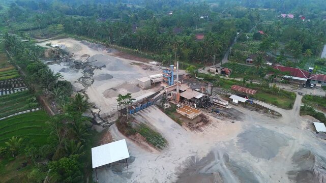 Aerial view of sand mining site with industrial machinery and piles of gravel surrounded by lush green vegetation and rice fields in Kecamatan Tempel, Daerah Istimewa Yogya, Indonesia.