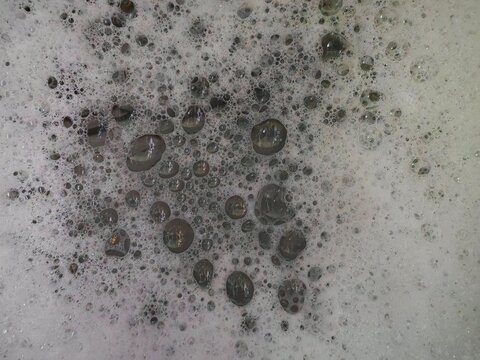 Macro close-up of grey soapy foam with scattered water droplets and bubbles, creating an abstract textured background pattern.