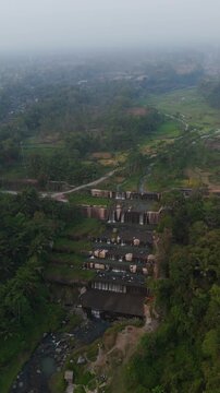 Aerial view of the tiered Grojogan Watu Purbo waterfall surrounded by lush tropical vegetation and rice fields in Kecamatan Tempel, Daerah Istimewa Yogya, Indonesia.