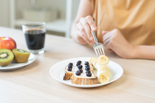 Closeup of Asian woman hand holding fork eating healthy breakfast toast with blueberry, banana, kiwi, red apple and black coffee on wooden table ai=t home, wellness and diet lifestyle concept.