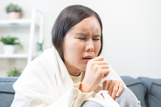 Sick Asian young woman coughing and closing mouth with hand, female wrapping in blanket having seasonal influenza or throat infection symptom while resting on sofa at home