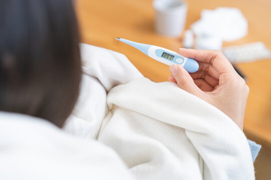 Close-up of sick Asian woman hand holding a digital thermometer showing high body temperature of 39.7 degrees celsius, female wrapping in white blanket having a severe fever at home