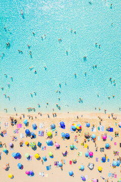 Aerial drone view of people relaxing on a white sand beach under colorful umbrellas. Bright sunny summer vacation scene by turquoise crystal clear sea water.