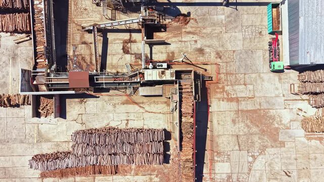 Workers are processing wood in a lumber yard. Machinery is moving logs while timber is stacked neatly on the ground. The scene shows a bustling work environment with equipment and logs.