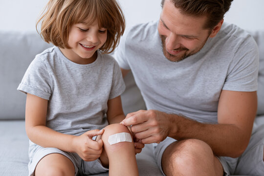 Man gently applies bandage to child's knee. Child smiles, looking at the bandage with delight. Father leans in, showing care and affection. Scene suggests healing, bonding, and tender moments