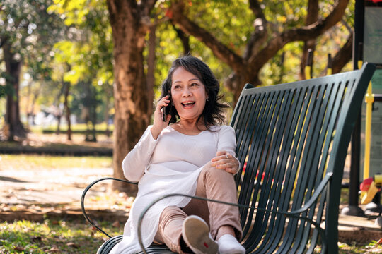 Older Asian Woman Talking on Phone Relaxing on Park Bench Happy Conversation in Outdoor Setting