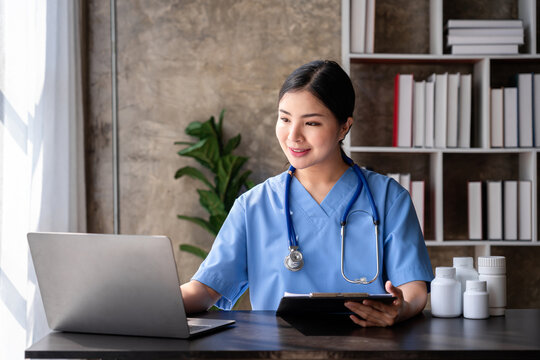Asian Women Doctor Using Laptop with Stethoscope Healthcare Professional in Blue Scrubs and Clipboard at Office Desk for Medical Research or Patient Consultation