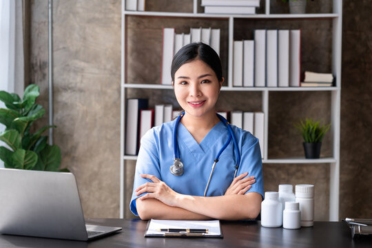 Asian Adult Woman Doctor With Stethoscope in Clinic Office Smiling With Laptop and Medicine Bottles on The Table for Healthcare