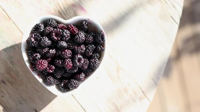 Heart shaped bowl of ripe blackberries glowing in sunlight on weathered wooden table, with shifting highlights, soft shadows, rich texture, natural color and rustic charm