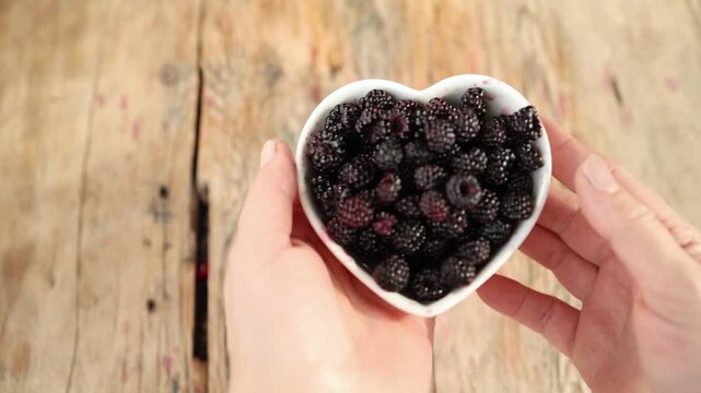 Hands presenting heart shaped bowl filled with ripe blackberries over rustic wooden table, glossy fruit, dark juice marks, warm natural light, close overhead composition