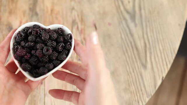Hands presenting heart shaped ceramic bowl filled with ripe blackberries on weathered wooden table under soft daylight, rustic summer food scene with rich color and natural texture