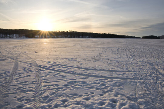 Winter sunset over frozen Lake arr in Dalsland, Sweden; golden sun rays shining behind an old stone ruin on a snowy shore with a peaceful, scenic wilderness atmosphere