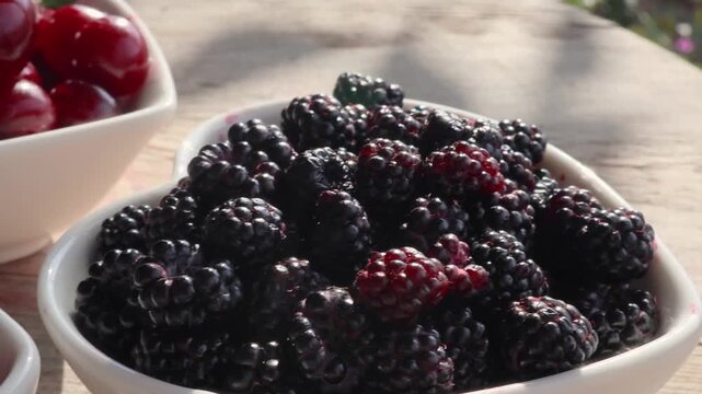 Close-up bowls filled with fresh blackberries, red currants, cherries, on weathered wooden table in warm sunlight, rustic outdoor food scene with rich color, texture, and natural summer harvest appeal