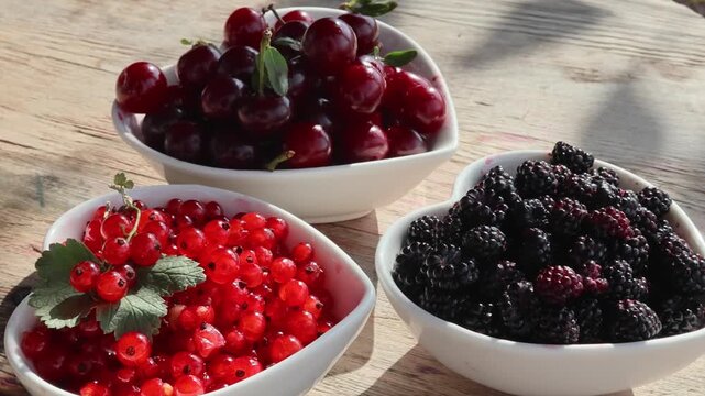 Close-up heart-shaped bowls of cherries, red currants and blackberries on weathered wooden table with changing sunlight, glossy highlights and rustic summer fruit arrangement