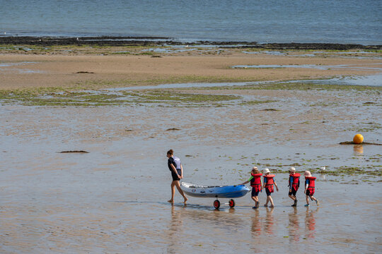 Langrune-Sur-Mer, France - 08 10 2026: An instructor woman pulling a kayak along the beach, followed by four young children wearing red life jackets and sea background