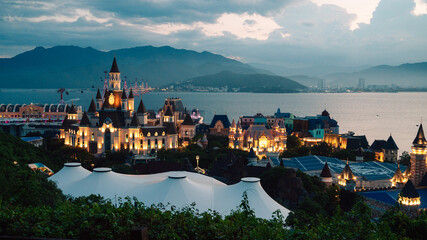 Illuminated castle-inspired of Vinpearl in VinWonders theme park overlook a scenic bay at dusk, creating a magical waterfront cityscape with mountains, dramatic clouds, and evening lights. © olinchuk
