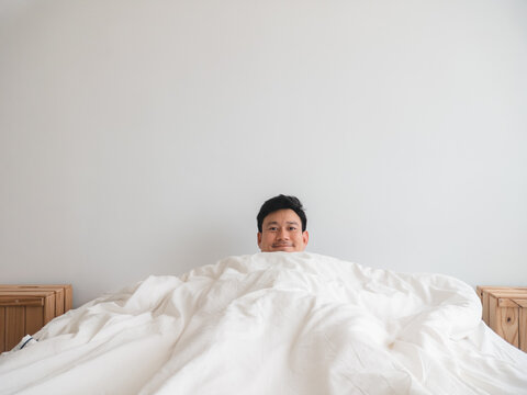 Relaxed Asian man smiling and peeking from under a white duvet in bed during morning.
