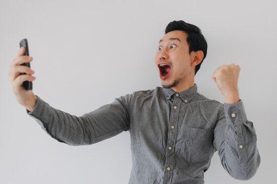 Excited Asian man celebrating success while looking at his smartphone with a triumphant fist.