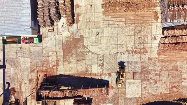 At a wood processing facility during daytime, workers manage logs and operate machinery on a large, open area. Stacks of wood are seen nearby, creating an organized scene.