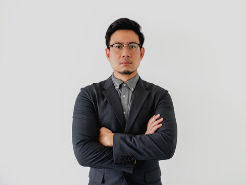 Serious Asian businessman in dark suit, glasses, arms crossed, white background