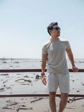 Young Asian Man in Sunglasses Standing by a Muddy Coastal Area on a Sunny Day