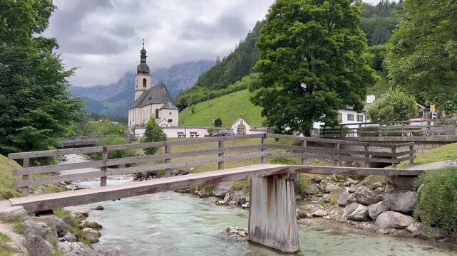 Motion of cars traffic in front of Parish Church of St. Sebastian (Pfarrkirche St. Sebastian Ramsau). Motion of mountain river, spring. Ramsau bei Berchtesgaden, Germany 