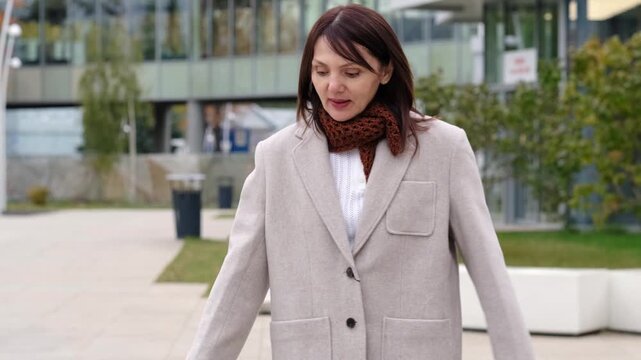 A medium outdoor shot shows a woman brushing her hand over a light wool blazer, feeling the natural fabric near a modern building. It suits slow fashion, autumn style, texture, and urban elegance.
