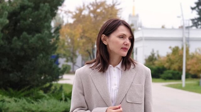 A soft outdoor portrait shows a thoughtful woman in a beige wool blazer looking down with calm emotion and autumn greenery behind. It suits mature beauty, urban style, wellness, and lifestyle stories.