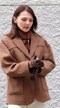 A stylish mature woman adjusts elegant black gloves beside a textured wall, dressed in layered autumn clothing with calm confidence. It suits fashion after fifty, urban elegance, and timeless style.
