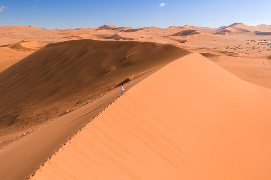 Aerial view shows a lone hiker walking a sharp sand dune ridge in Sossusvlei near Deadvlei, Namibia, with apricot dunes and dark wind shadow in bright daylight.