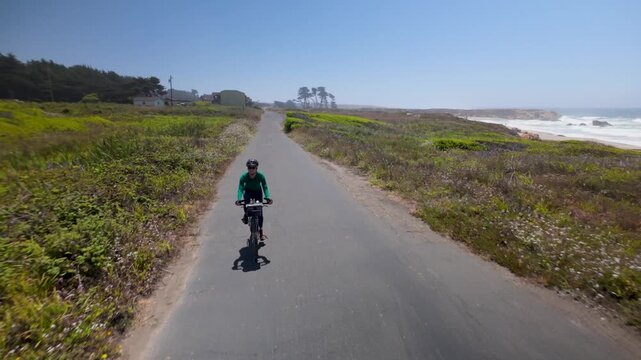 A woman rides her bike on the paved Coastal Trail in Fort Bragg, California with the ocean and flowers all around her.