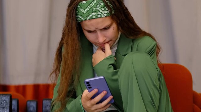 Worried high school girl with green bandana hugs her knees and anxiously reads chat on purple smartphone, afraid of new insults in group chat, theme cyberbullying and social media pressure.
