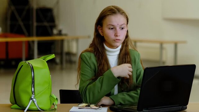 Young woman in green suit at large table feels overwhelmed by work tasks on laptop, holding her face in hands, concept workplace stress and overload for generation z.