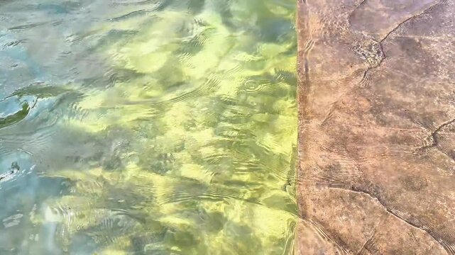 Gentle water surface rippling with sunlight reflections over a stone poolside pavement, creating a serene and abstract summer vacation background concept