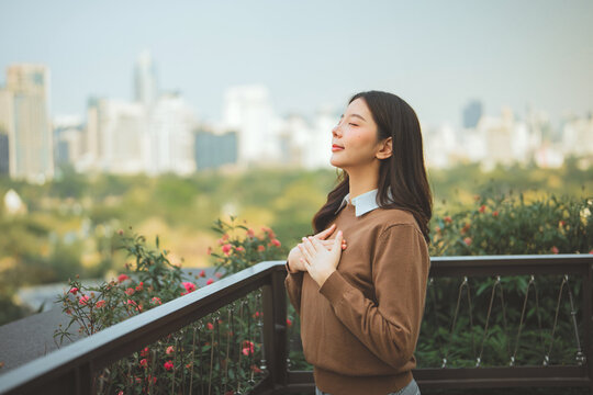 Asian woman holding hands on chest closed eyes taking deep breath at rooftop balcony, peaceful serene female enjoying fresh air and city park view for mental health, mindfulness, self care and wellnes