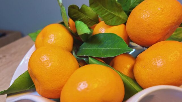 Fresh ripe mandarins with green leaves piled in white curved ceramic bowl on wooden table.