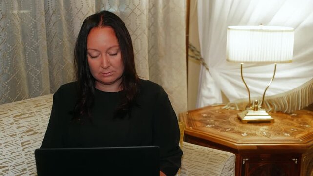 Professional woman with long dark hair concentrates on laptop work in upscale hotel suite decorated with wooden furniture, brass lamp, patterned curtains, bathed in natural light.
