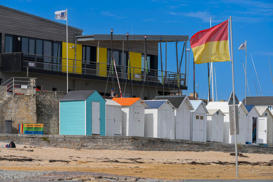 Langrune-Sur-Mer, France - 08 10 2026: View graphical colorful wooden beach huts along the stone jetty, sand beach, a yellow and red flag and blue sky behind
