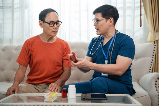 Male Nurse Explaining a Medicine Bottle to an Asian Elderly Man during a Senior Medication Consultation with Pill Organizer and Digital Tablet at Home