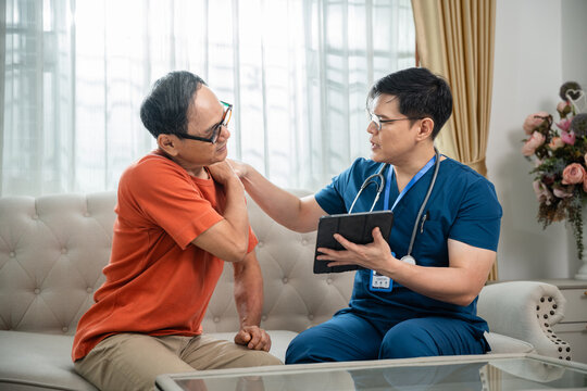 Male Nurse Assessing Shoulder Pain in an Asian Elderly Man while Using a Digital Tablet during a Home Healthcare Consultation in a Living Room